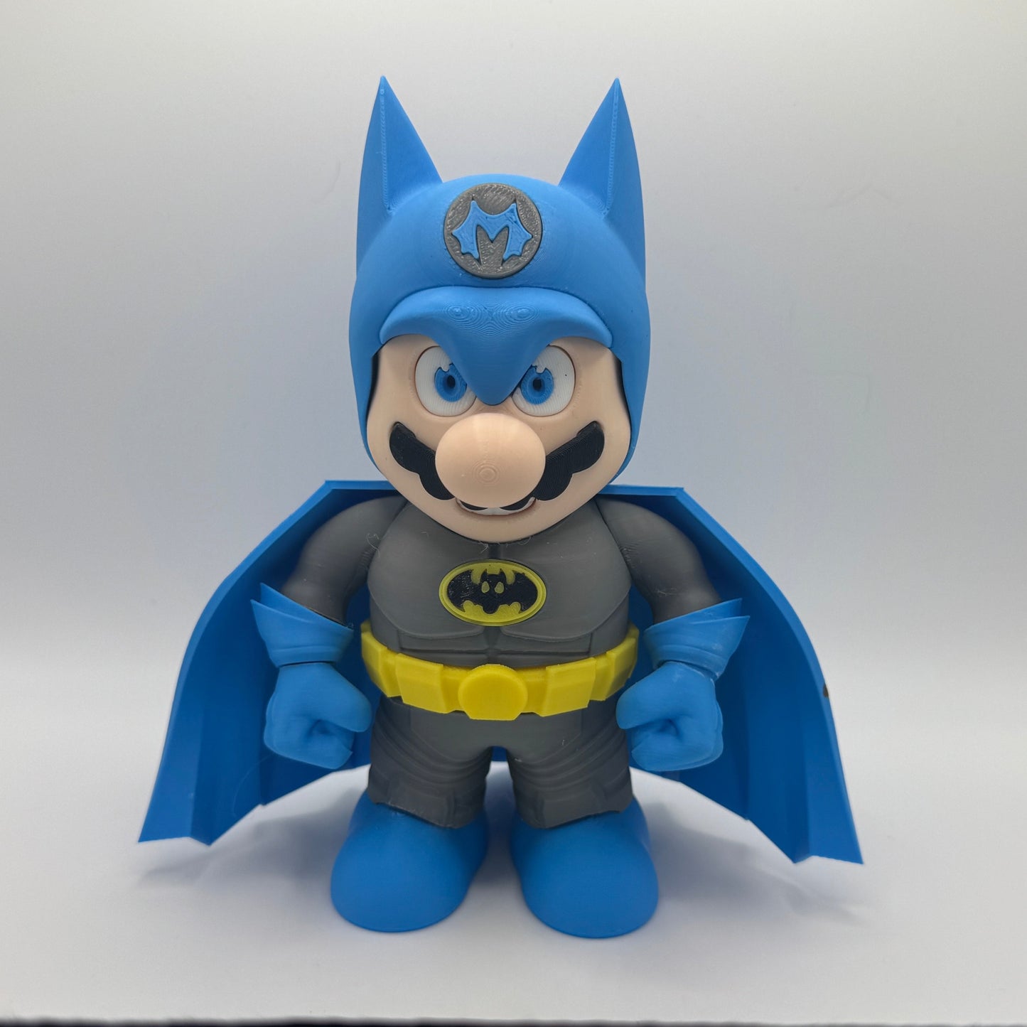 Toy figure of a Mario character wearing a blue cape and gray outfit with a Batman logo on a white background